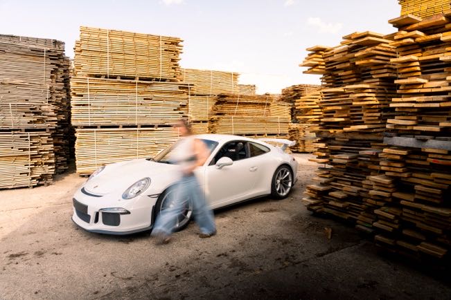 Voiture de sport blanche dans une cour de scierie, entre des piles de bois